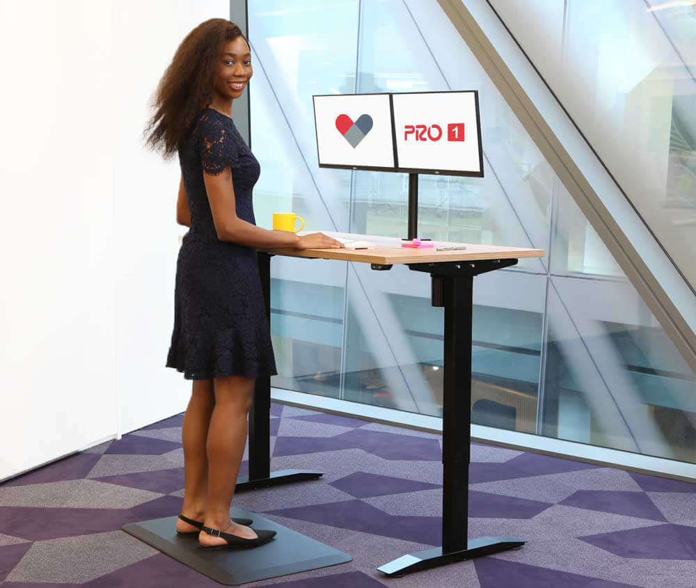 Woman using PRO 1 single motor electric standing desk with dual monitors in a modern office setting.