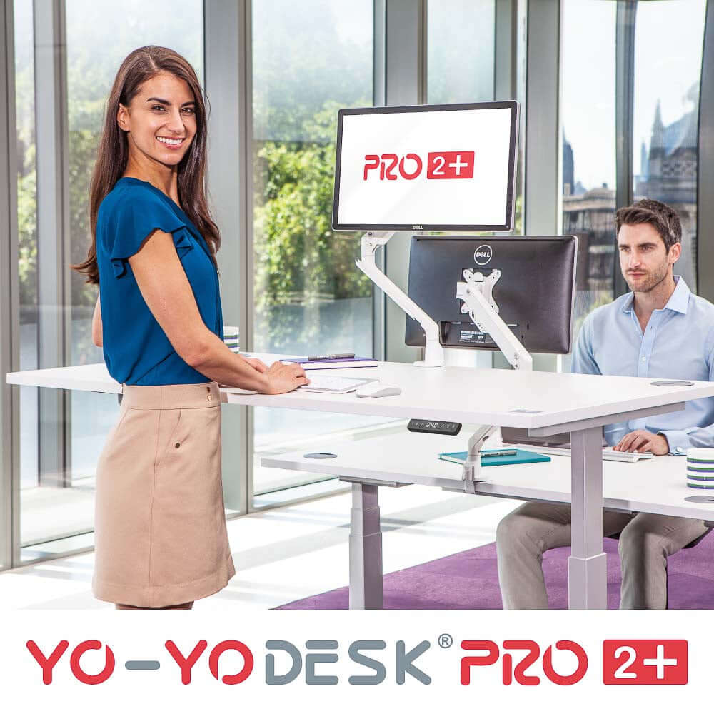 Woman using PRO 2+ dual motor electric standing desk with dual monitors in a modern office setting.